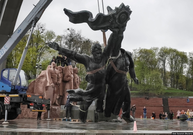 Photograph showing a large statue of two men being lowered by a crane. A small crowd watches in the background.